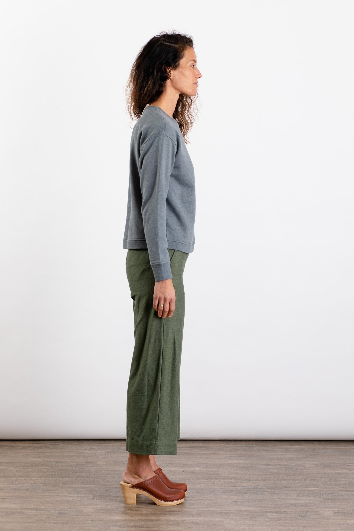 A woman stands in profile against a plain white background, wearing the Bridge & Burn Hawthorne Boxy Crew Sweatshirt in Storm Blue, paired with olive green wide-leg pants and brown wooden-heeled clogs.