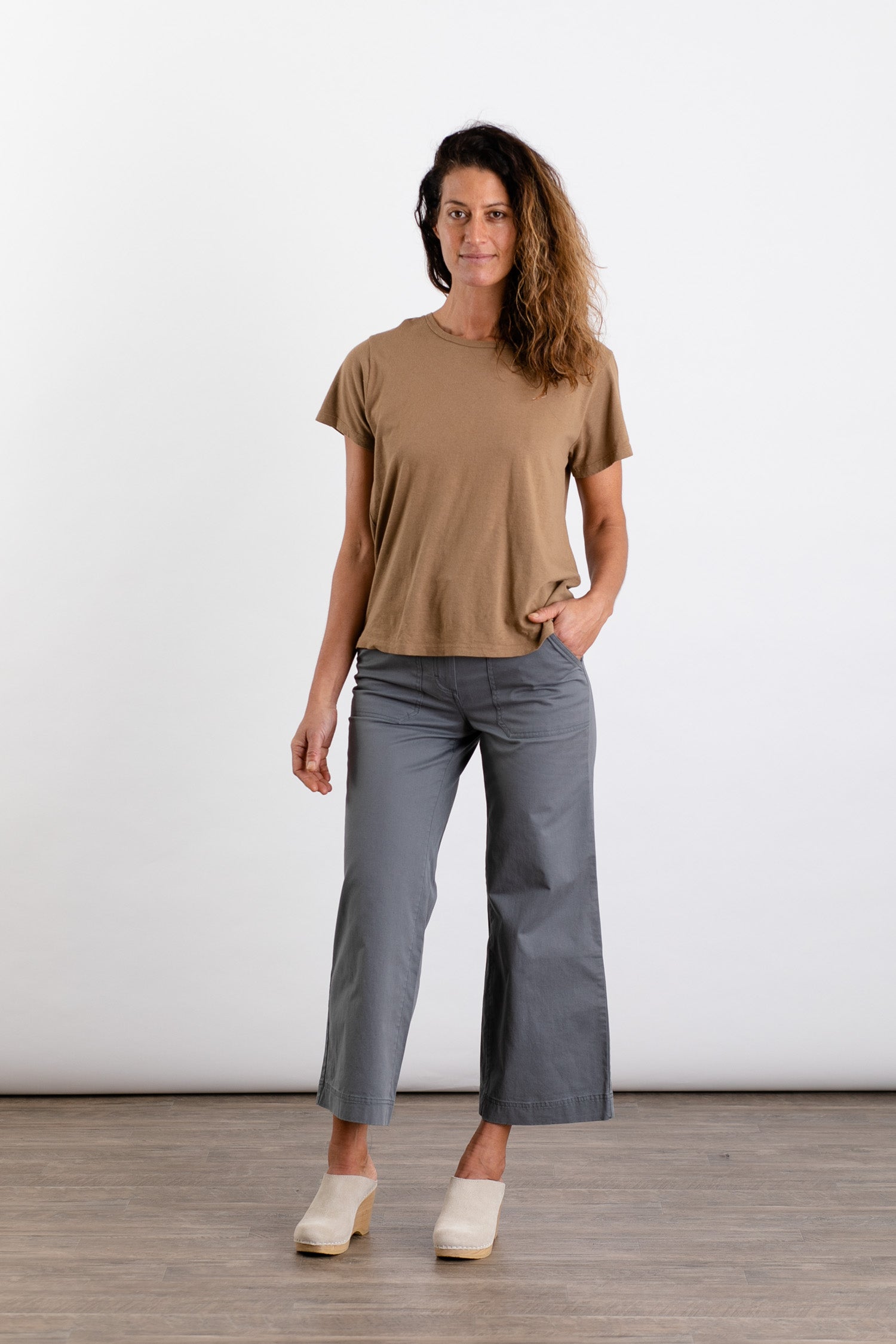 A woman stands against a white background, smiling slightly with one hand in her pocket. She wears the Jungmaven Ojai Tee in Coyote—a relaxed fit crewneck—paired with gray wide-leg pants and light-colored clogs.