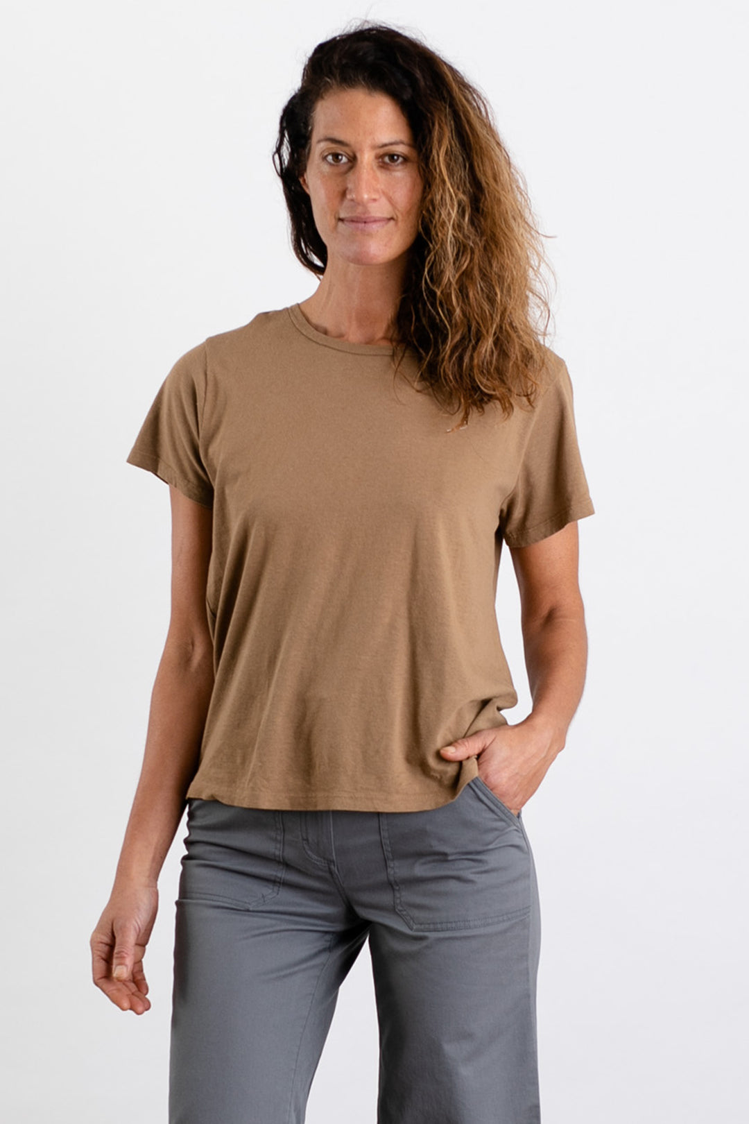 A woman with wavy brown hair wears the Jungmaven Ojai Tee in Coyote with gray pants, standing against a plain white background, one hand in her pocket and a relaxed expression.