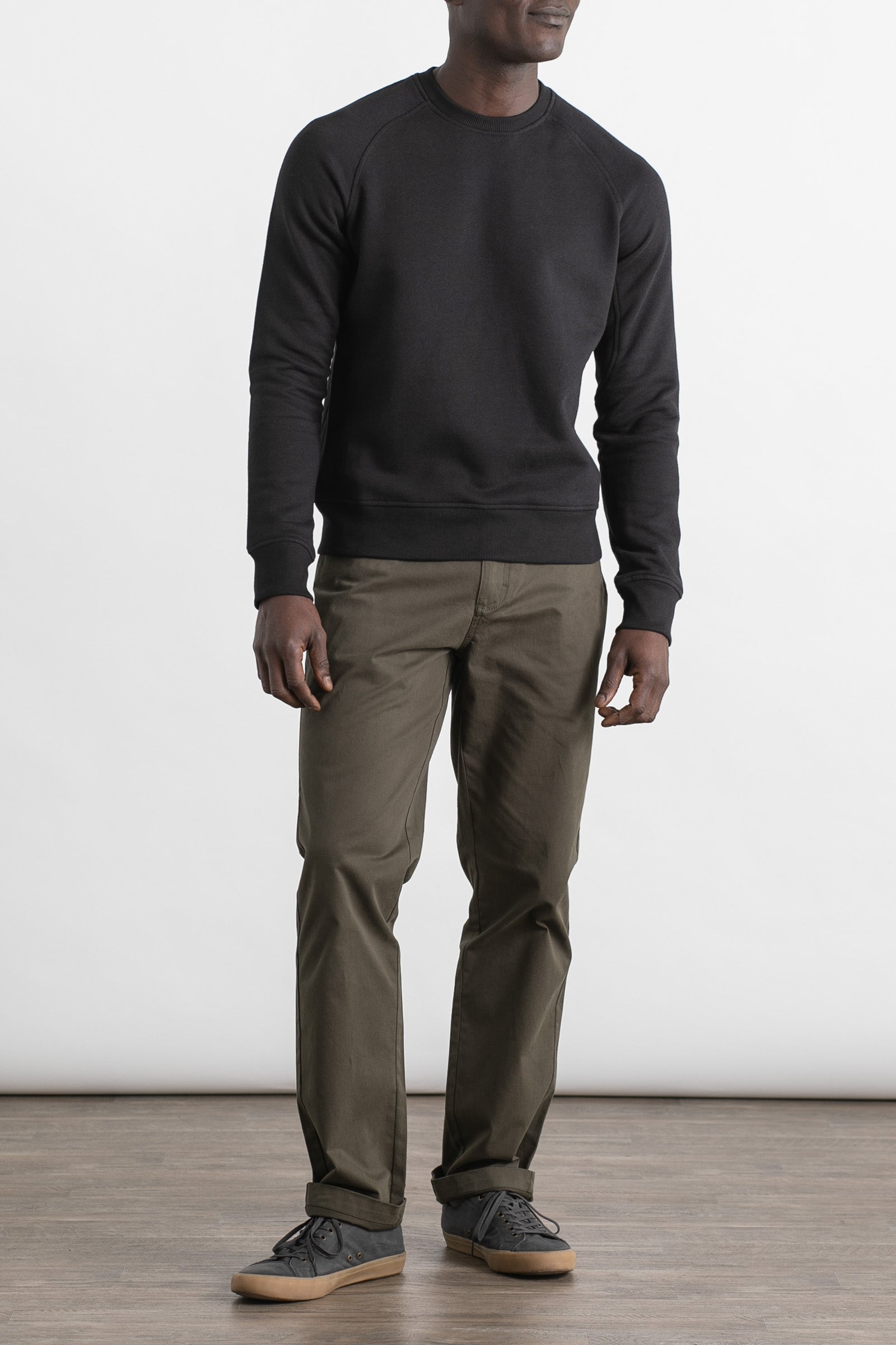 A man stands indoors against a plain white background, wearing a Bridge & Burn Fremont Crew Sweatshirt in black, olive green pants, and gray sneakers. His face is not visible.