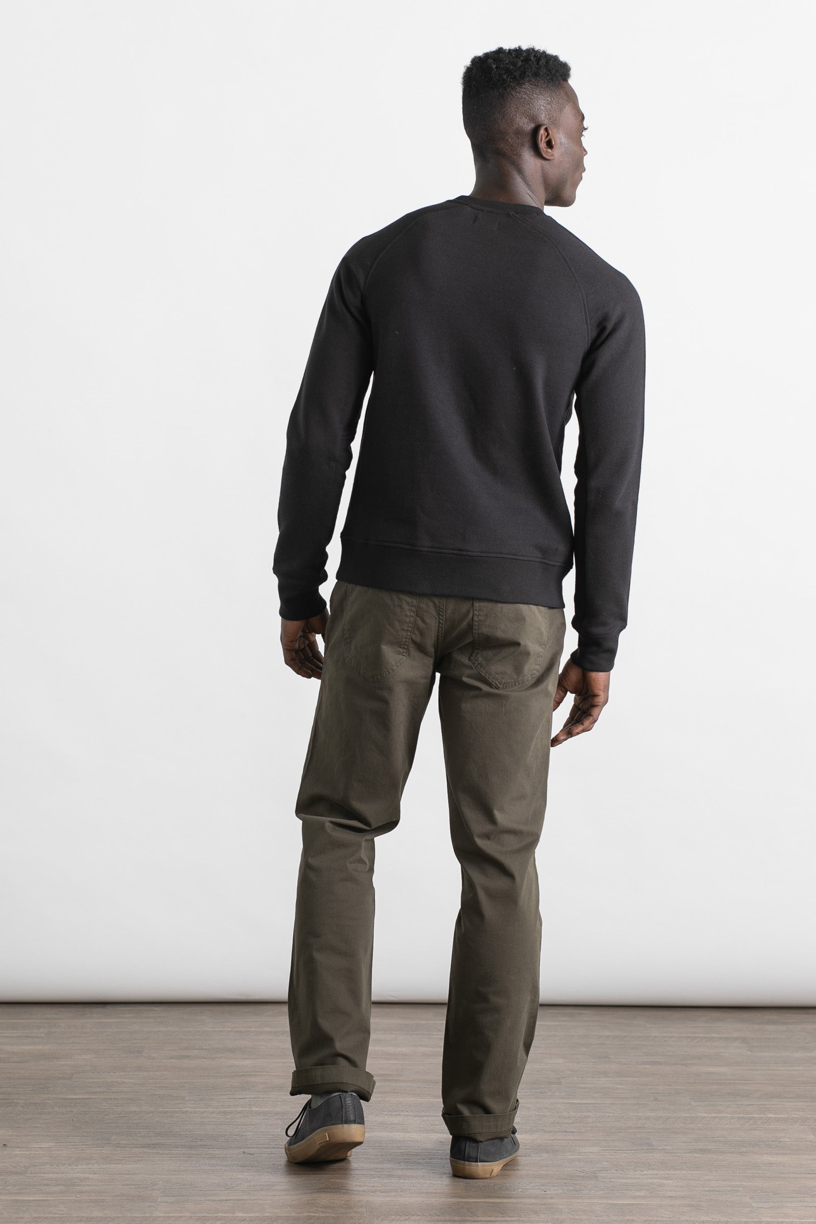 A man stands indoors on a wooden floor facing away from the camera, wearing the Bridge & Burn Fremont Crew Sweatshirt in black, olive green pants, and black sneakers. The background is plain white.