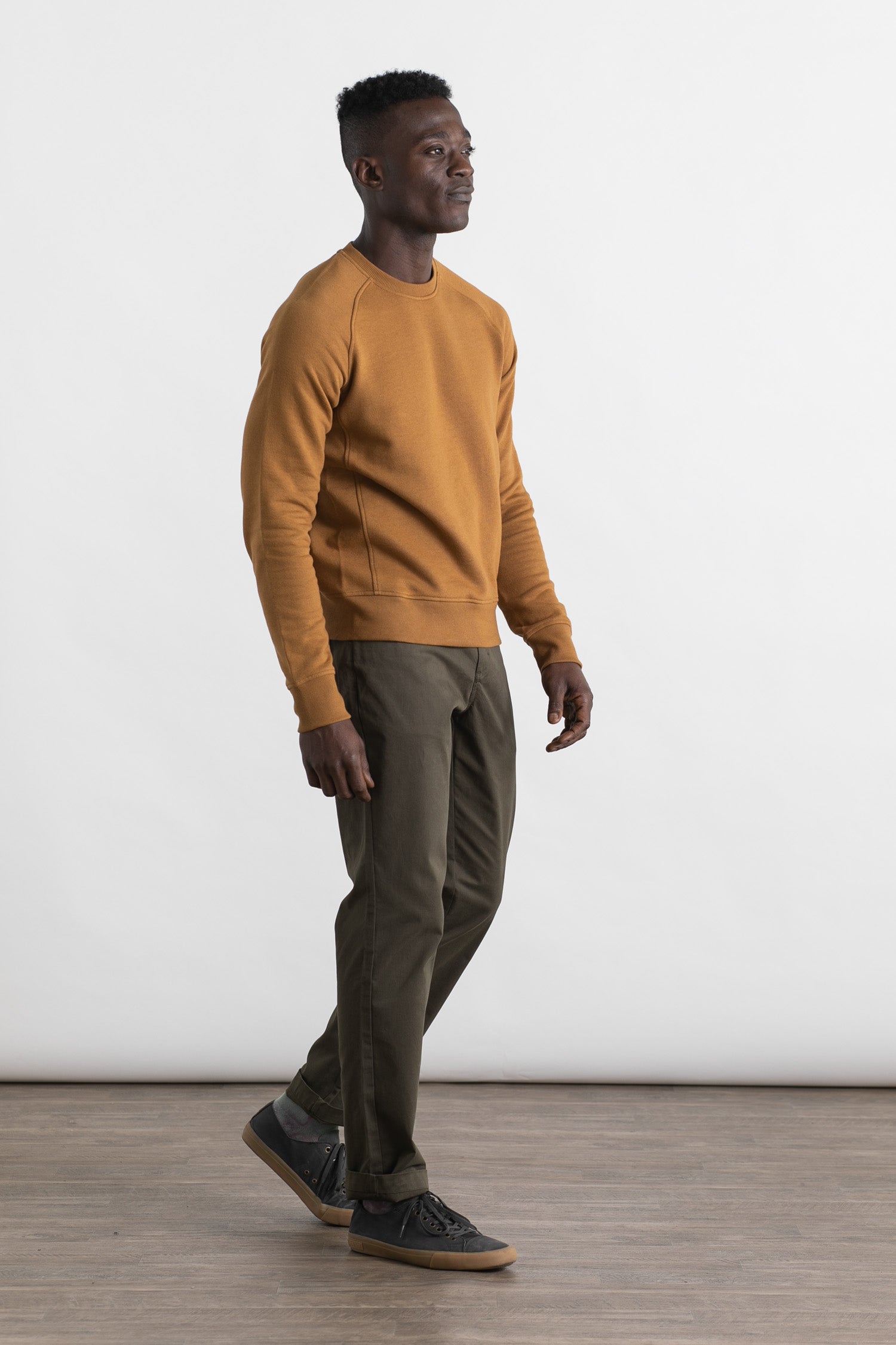 A man wears the Bridge & Burn Fremont Crew Sweatshirt in bronze, made from organic cotton, styled with olive green pants and dark sneakers, walking on a wooden floor against a plain white background.