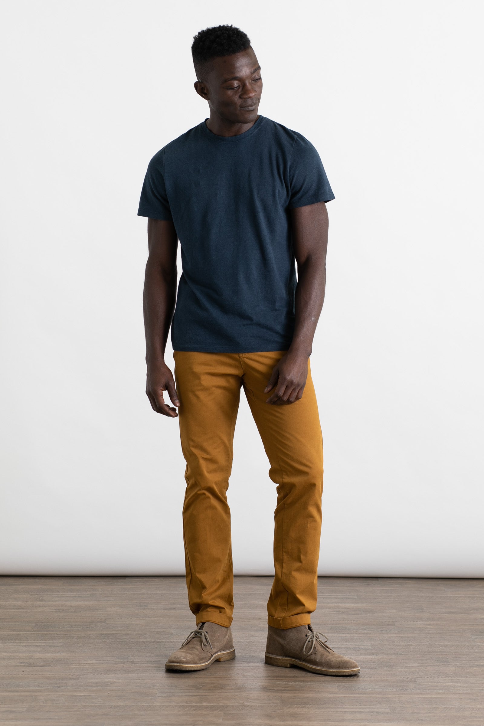 A man stands on a wooden floor against a white background, wearing a dark blue t-shirt and Bridge & Burn Bradley Slim Pant in Flax. The 5-pocket style of these pants adds a modern touch to his relaxed look.