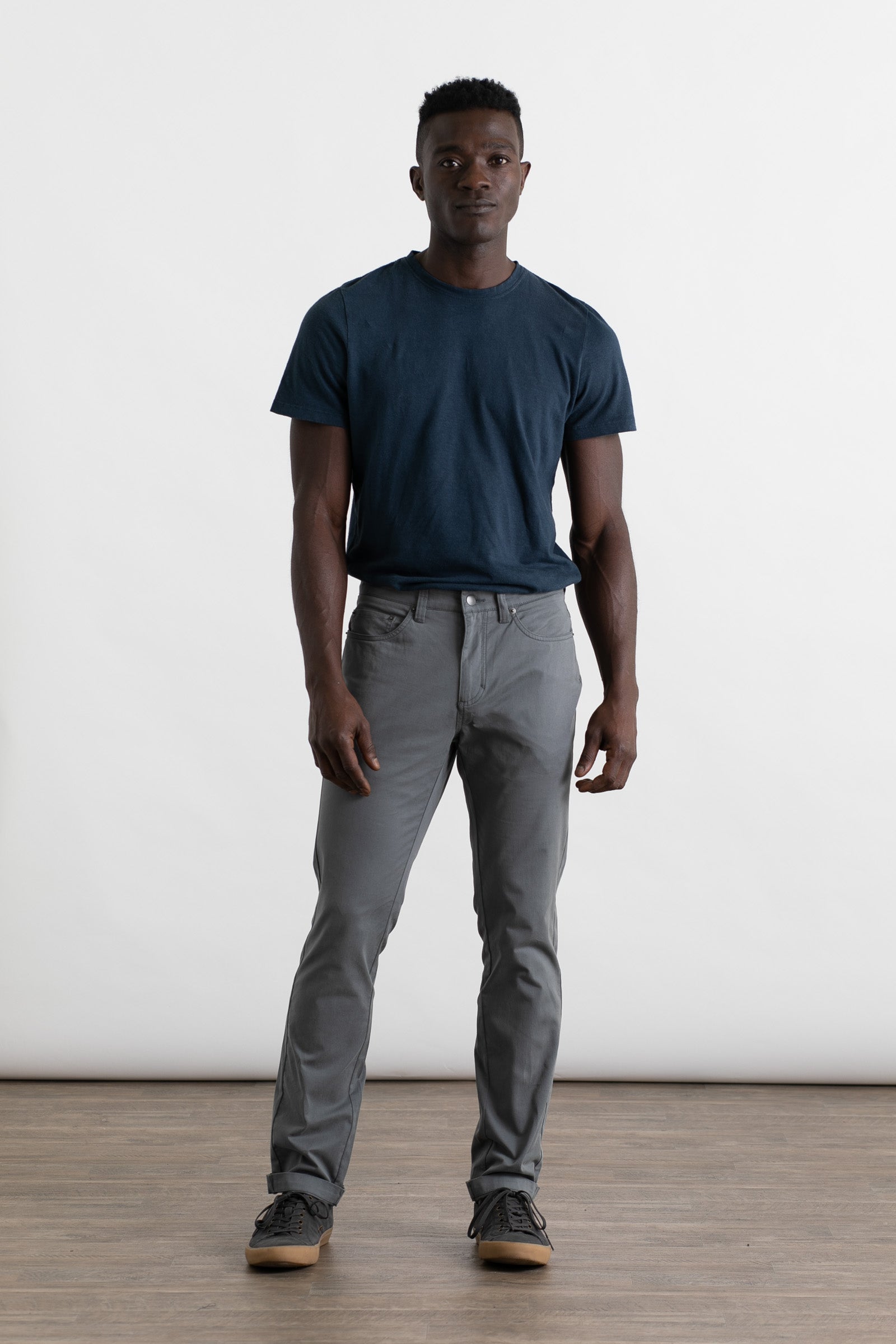A man stands on a wooden floor against a plain white background, wearing a dark blue t-shirt and Bridge & Burn’s Bradley Slim Pant in Storm Blue, featuring a 5-pocket style. He faces forward with a neutral expression and relaxed posture.