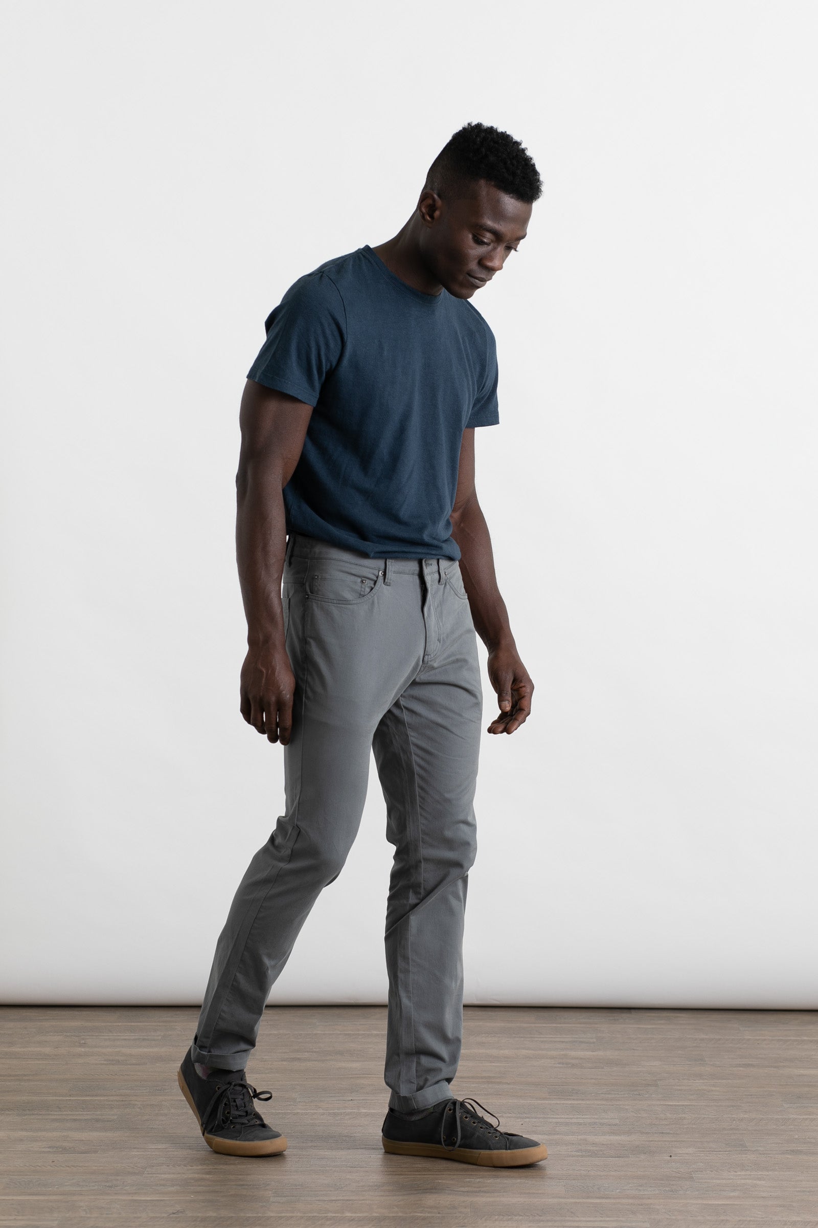 A man wearing Bridge & Burn's Bradley Slim Pant in Storm Blue walks indoors on a wooden floor against a plain white background, looking slightly downward.