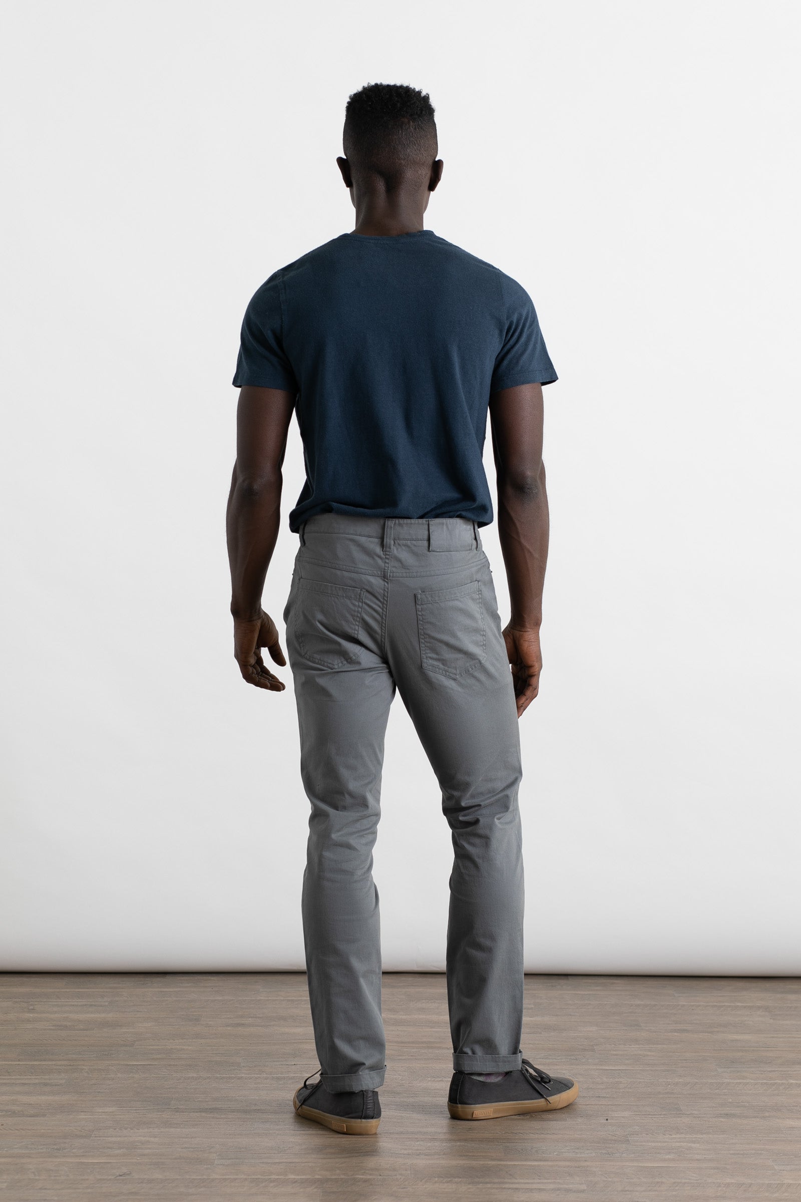 A man faces away from the camera wearing Bridge & Burn's Bradley Slim Pant in Storm Blue, a navy blue t-shirt, and dark sneakers. He stands on a wooden floor against a plain white background.
