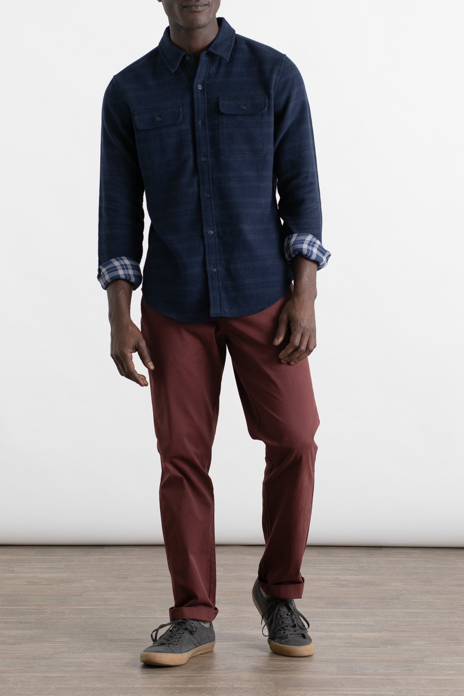 A person wearing a navy button-up shirt, Bridge & Burn Polk Pant in burgundy, and black sneakers stands on a wooden floor against a plain white background.