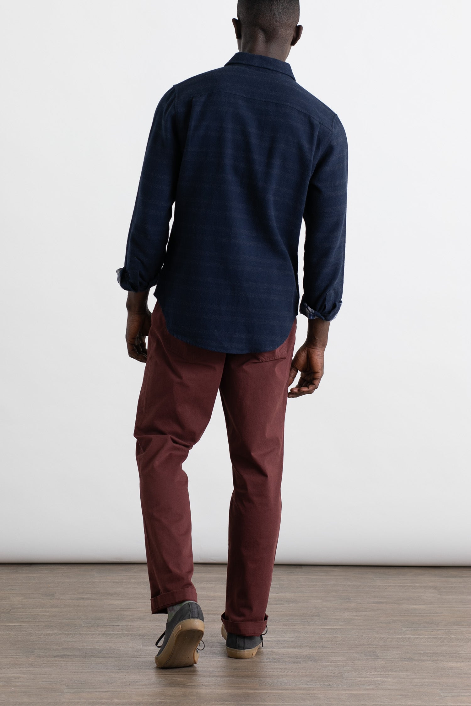 A person faces away from the camera, wearing Bridge & Burn's Polk Pant in Burgundy—a classic 5-pocket organic cotton style—with a dark blue long-sleeve shirt and gray shoes, standing on a wooden floor against a white background.