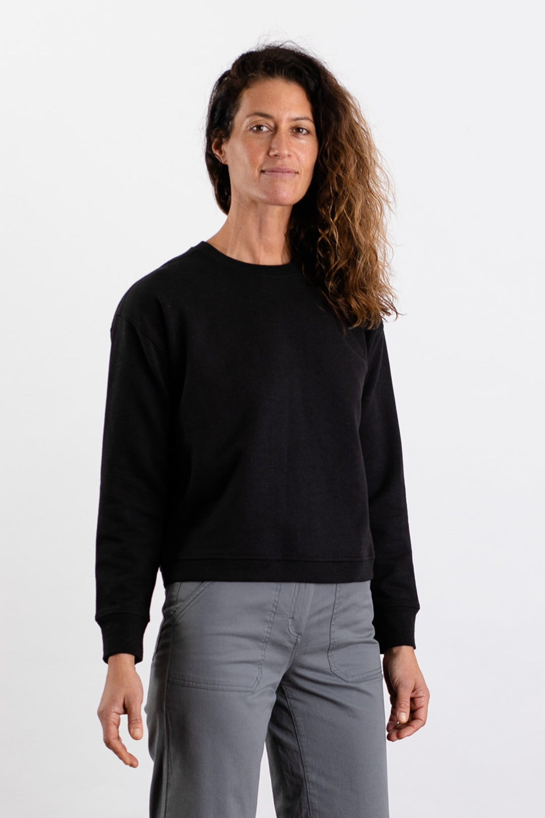 A person with long, wavy brown hair faces the camera, wearing the Bridge & Burn Hawthorne Boxy Crew Sweatshirt in black and gray pants against a plain white background.