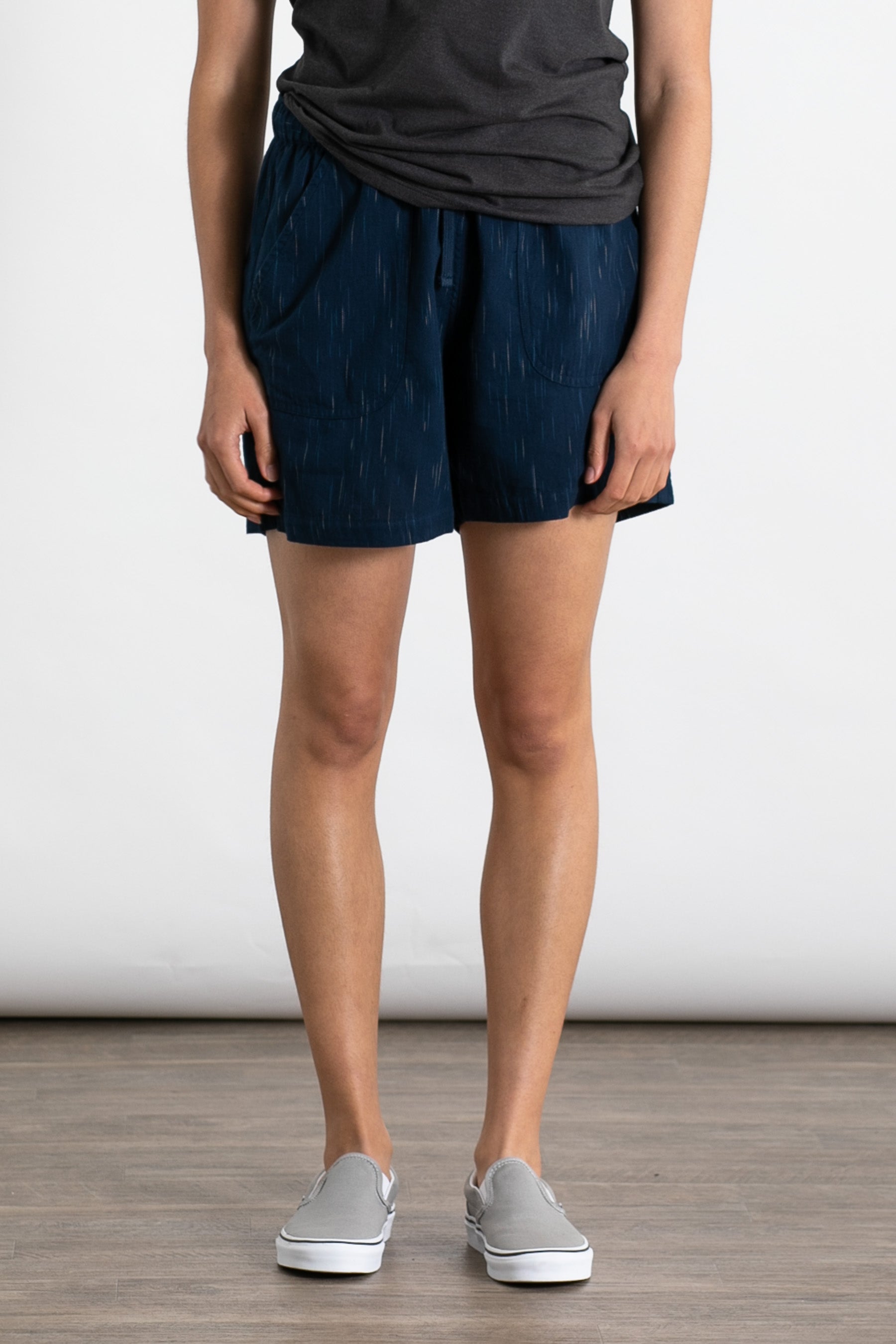 A person indoors wears the Bridge & Burn Nico Short in Navy Space Dye, a dark gray t-shirt, and gray slip-on shoes, standing against a plain white background with a wooden floor.