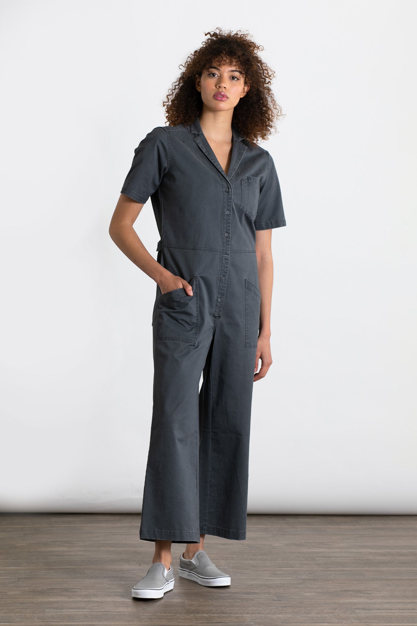 A person with curly hair wears the Bridge & Burn Lou Utility Jumpsuit in Slate—short-sleeve, wide-leg, adjustable buttons, and front pockets—with light gray slip-on shoes, standing indoors on a wooden floor against a white background.