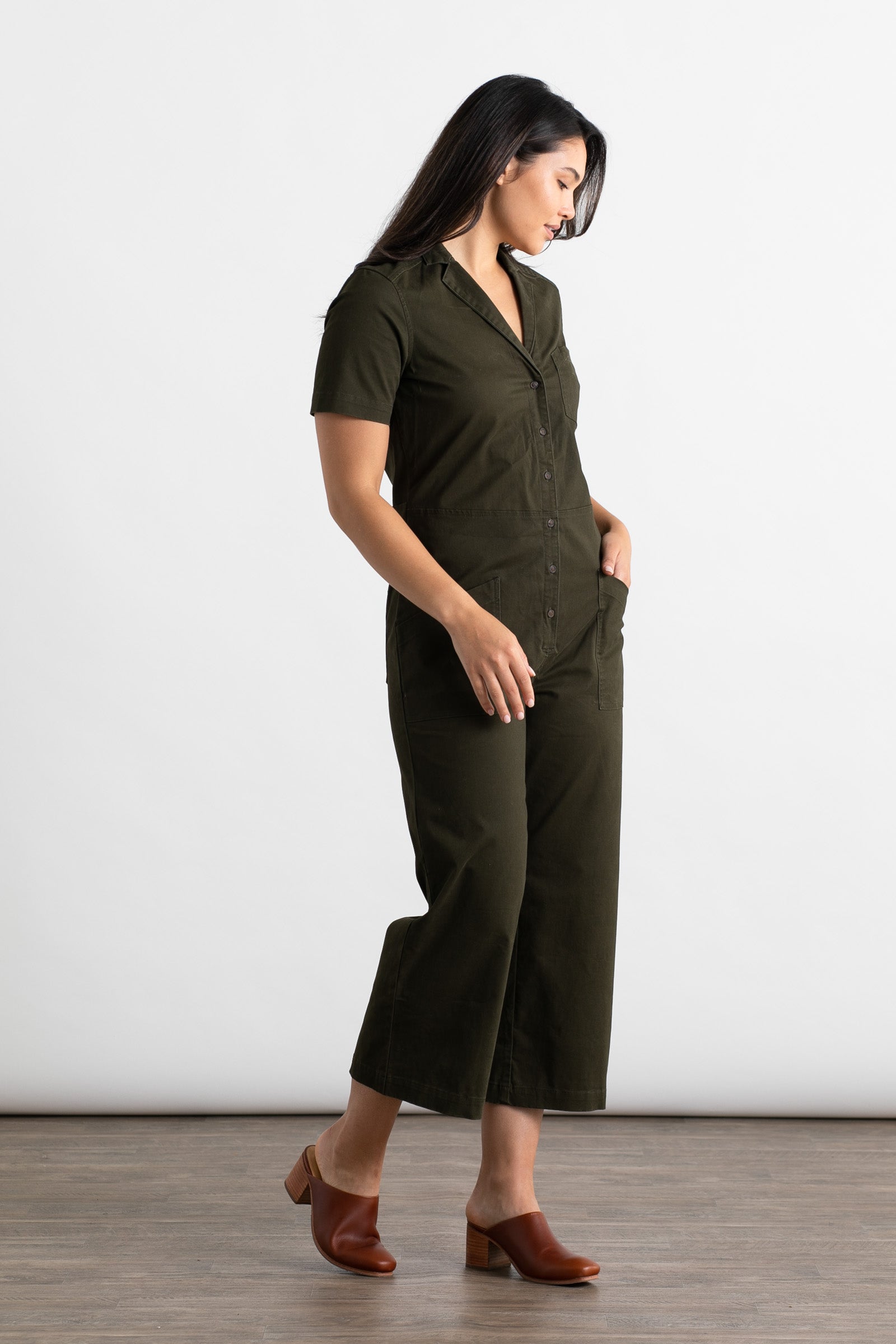 A woman with dark straight hair stands sideways against a white background, wearing the Bridge & Burn Lou Utility Jumpsuit in Olive with short sleeves and cropped wide legs, paired with brown heeled mules and one hand in her pocket.