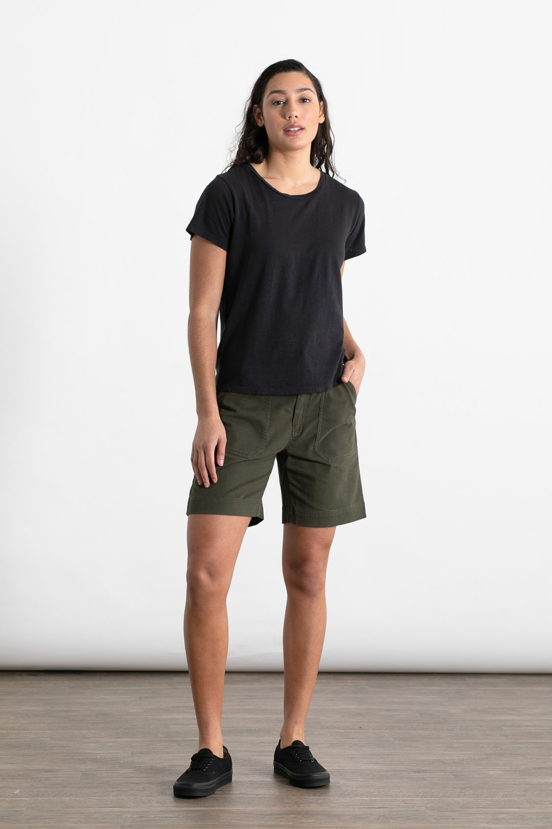 A person stands indoors against a plain white background, wearing the Jungmaven Ojai Tee in black, olive green shorts, and black sneakers, with one hand in their pocket and a neutral expression.