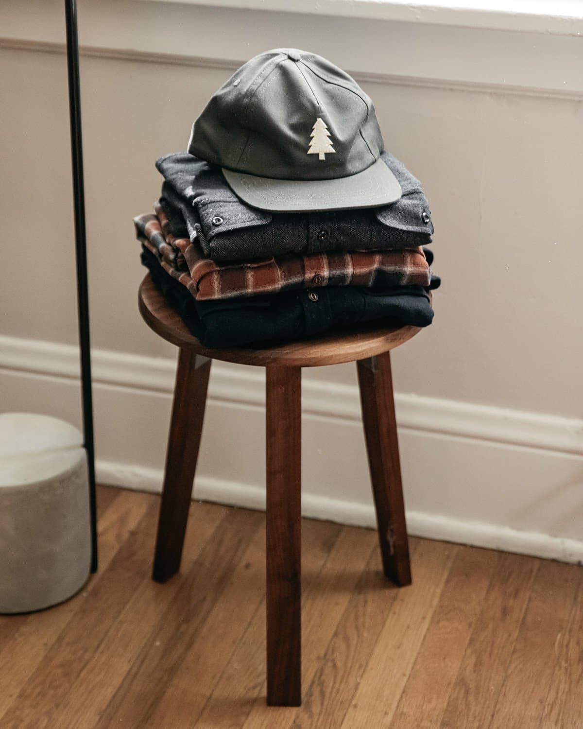 A stack of folded clothes topped with the Bridge & Burn Tree Cap in Olivewood, featuring a white tree logo, sits on a wooden stool in a room with wooden floors and white walls.
